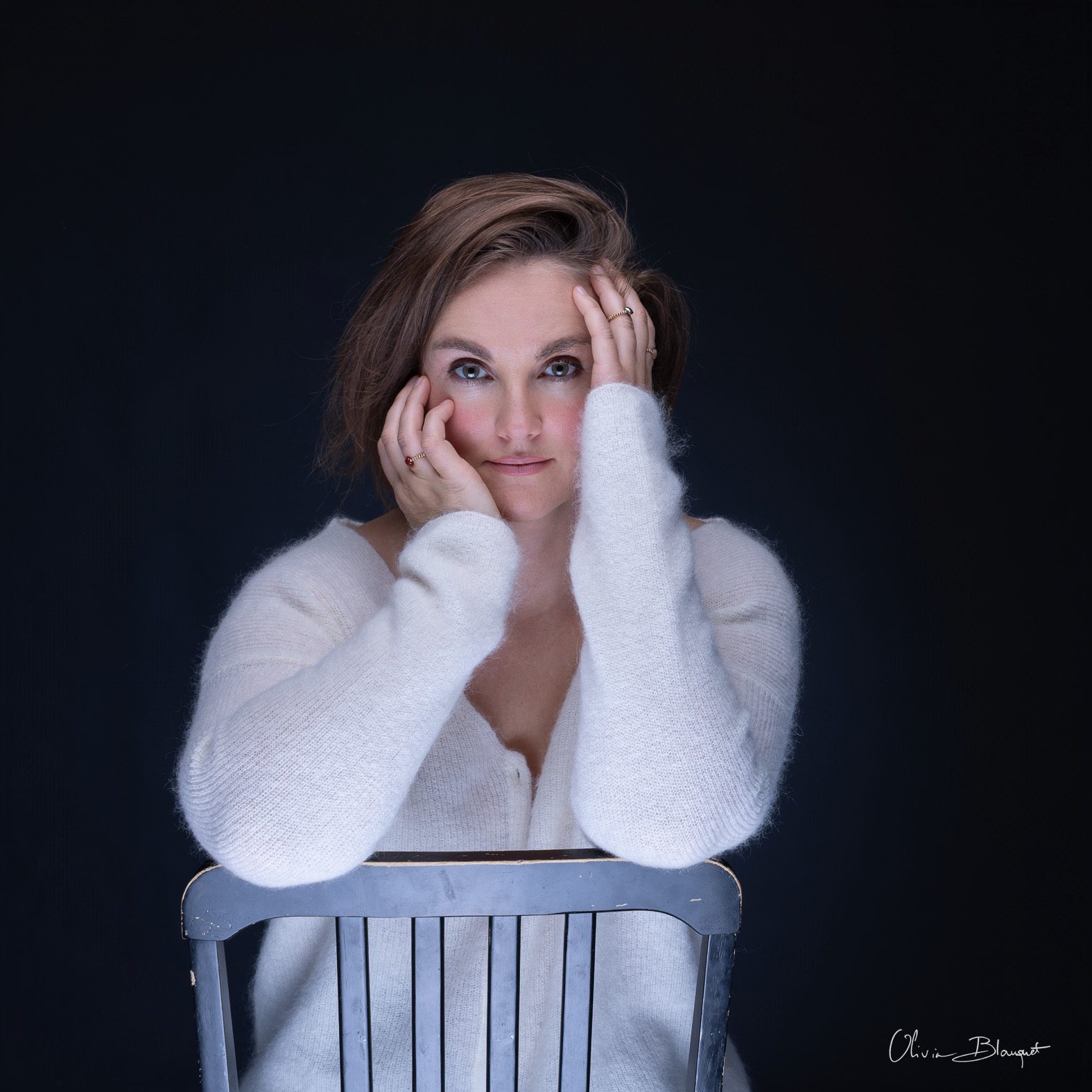 Portrait artistique d'une femme en pull blanc doux, mains sur le visage et regard intense, séance estime de soi par Olivia Blanquet.